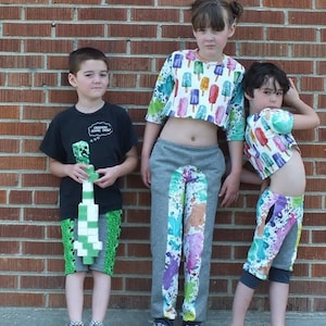 May include: Three children wearing colorful clothing with graphic prints. The child on the left is wearing a black t-shirt with a green and white graphic print and green and black pants. The child in the middle is wearing a grey crop top with a white and pink popsicle print and grey pants with a white, pink, and purple paint splatter design. The child on the right is wearing a white and blue crop top with a popsicle print and white and blue pants with a paint splatter design.