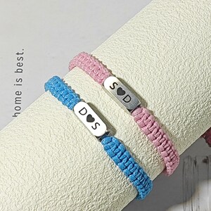 May include: Two braided bracelets, one blue and one pink, with silver rectangular charms. The blue bracelet has the letters "D S" with a heart symbol, and the pink bracelet has "S D" with a heart. The text "home is best" is visible.