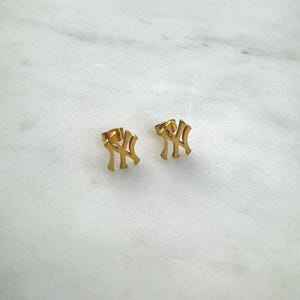 May include: A pair of gold-colored stud earrings. Each earring features the interlocking "NY" logo, a symbol of the New York Yankees baseball team. The earrings are displayed on a white marble surface.