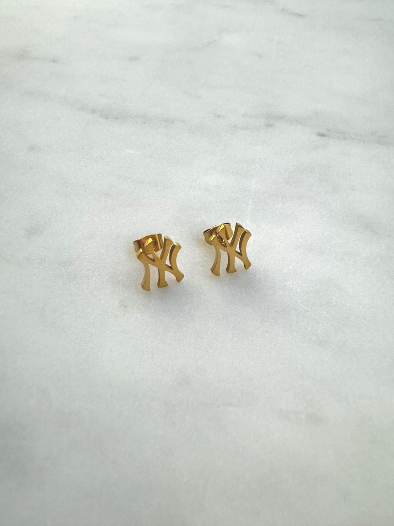 May include: A pair of gold-colored stud earrings. Each earring features the interlocking "NY" logo, a symbol of the New York Yankees baseball team. The earrings are displayed on a white marble surface.