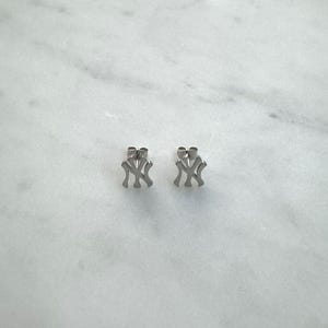 May include: A pair of silver-colored stud earrings, each featuring the interlocking "NY" logo. The earrings are displayed on a white marble surface, showcasing their simple, yet recognizable design. These earrings are a classic accessory.