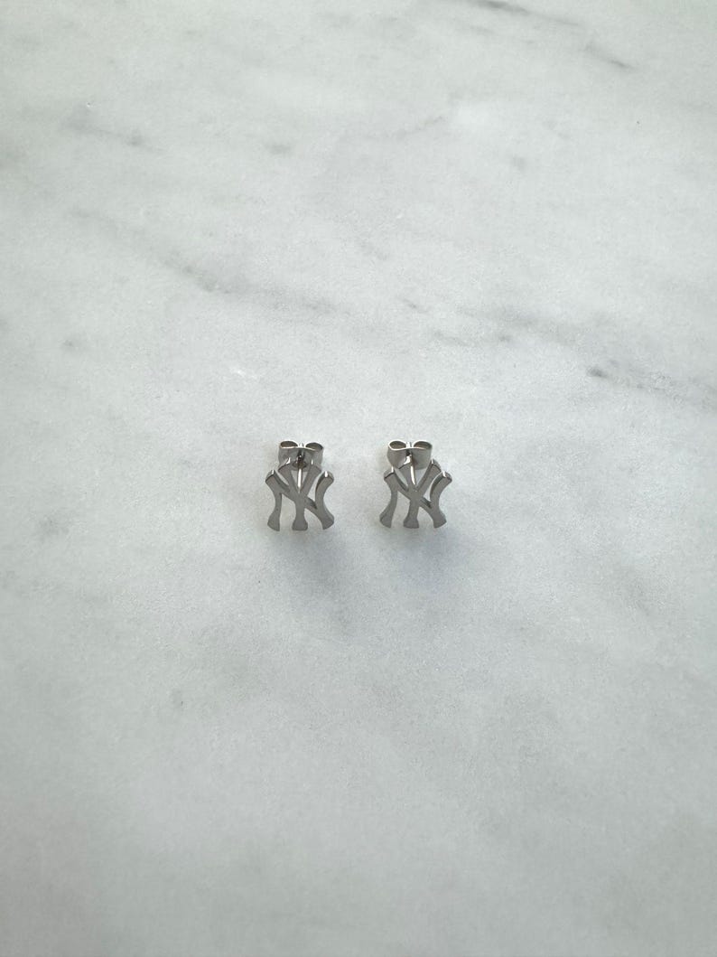 May include: A pair of silver-colored stud earrings, each featuring the interlocking "NY" logo. The earrings are displayed on a white marble surface, showcasing their simple, yet recognizable design. These earrings are a classic accessory.