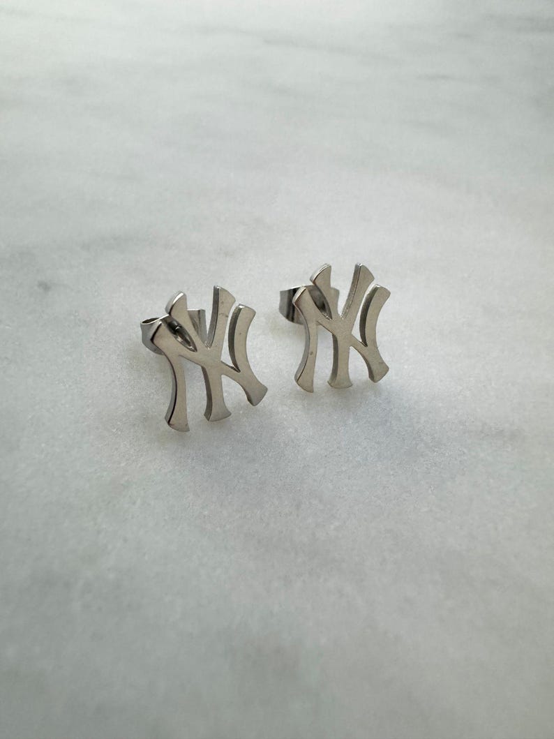 May include: A pair of silver-colored stud earrings. Each earring features the stylized interlocking "NY" logo, a symbol of the New York Yankees baseball team. The earrings are set against a light, marble-like background.