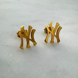 May include: A pair of gold-colored stud earrings. Each earring features the interlocking "NY" logo, a symbol of the New York Yankees baseball team. The earrings are set against a white and gray marble background.