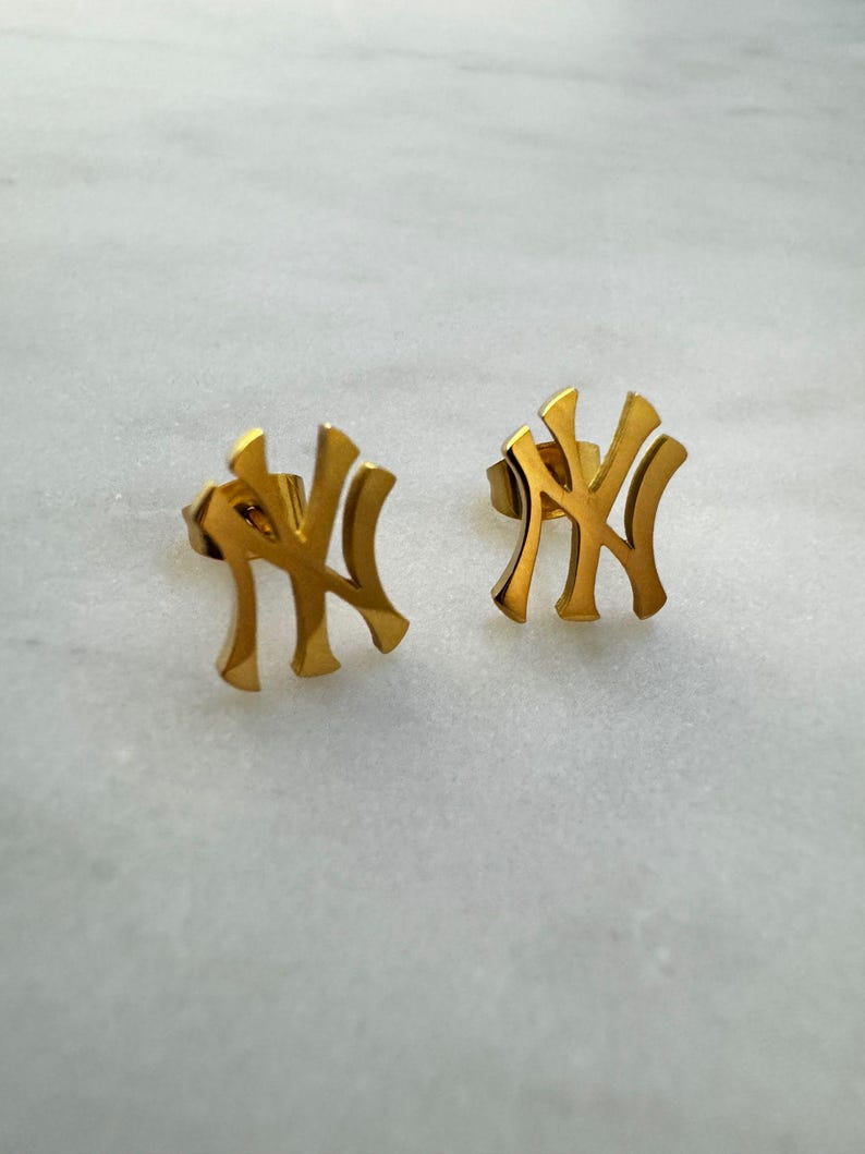 May include: A pair of gold-colored stud earrings. Each earring features the interlocking "NY" logo, a symbol of the New York Yankees baseball team. The earrings are set against a white and gray marble background.