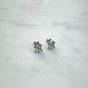 May include: A pair of silver-colored stud earrings. Each earring features the interlocking "NY" logo, a symbol of the New York Yankees baseball team. The earrings are displayed on a white marble surface.