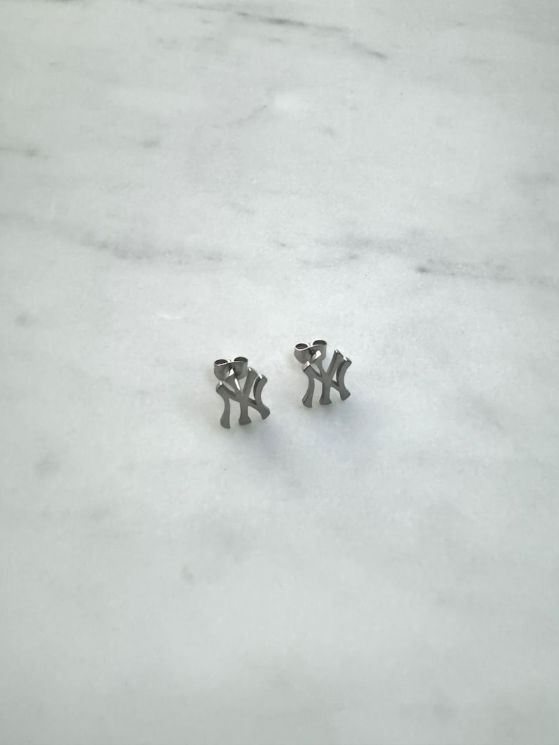 May include: A pair of silver-colored stud earrings. Each earring features the interlocking "NY" logo, a symbol of the New York Yankees baseball team. The earrings are displayed on a white marble surface.
