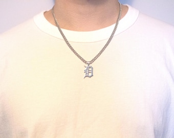 May include: Silver-toned chain necklace with a pendant featuring the letter "D" in an ornate, gothic-style font. The necklace is worn against a white t-shirt.