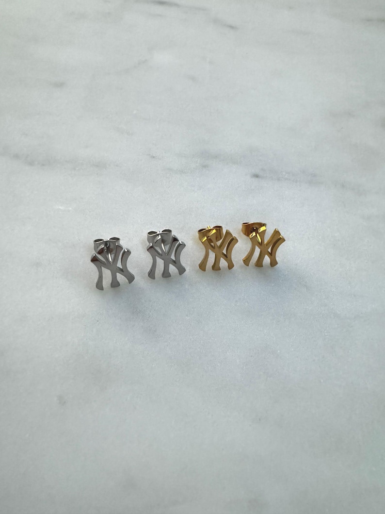 May include: Two pairs of stud earrings. One pair is silver-toned, and the other is gold-toned. Each earring features the interlocking "NY" logo, a symbol of the New York Yankees baseball team.