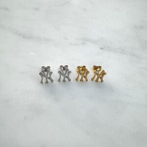 May include: Four stud earrings in a row, each featuring the interlocking "NY" logo. Two pairs are silver-toned, and the other two are gold-toned. The earrings are displayed on a white marble surface.
