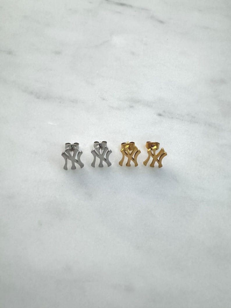 May include: Four stud earrings in a row, each featuring the interlocking "NY" logo. Two pairs are silver-toned, and the other two are gold-toned. The earrings are displayed on a white marble surface.