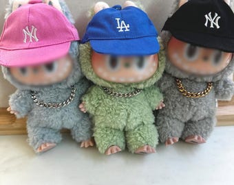 May include: Three plush figures in blue, green, and gray, each wearing a baseball cap and a gold chain necklace. The caps are pink, blue, and black, with white logos. The figures have a furry texture.