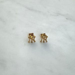 May include: A pair of gold-colored stud earrings. Each earring features the interlocking "NY" logo, a recognizable symbol. The earrings are small and appear to be made of metal. The background is a light-colored, marble-like surface.