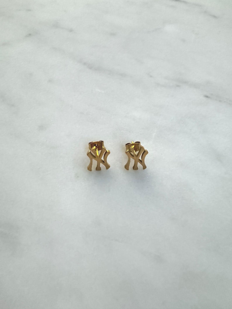May include: A pair of gold-colored stud earrings. Each earring features the interlocking "NY" logo, a recognizable symbol. The earrings are small and appear to be made of metal. The background is a light-colored, marble-like surface.