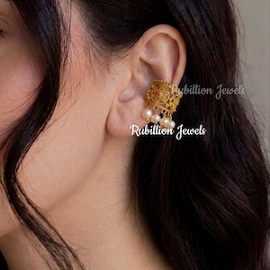 Puede incluir: Un pendiente tipo ear cuff dorado con un intrincado patrón de filigrana y tres perlas blancas colgantes. La pieza de joyería tiene grabado el texto Rubillion Jewels, que aparece varias veces sobre la superficie del diseño metálico.
