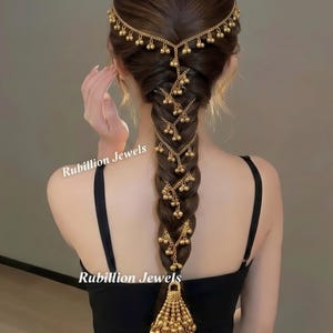 May include: A gold-toned hair accessory featuring a delicate chain headpiece that wraps around the crown and cascades down a braided hairstyle. The piece is adorned with numerous small, dangling gold spheres and a large, ornate tassel at the base.