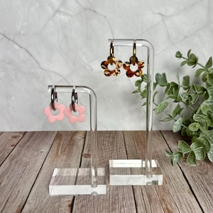 May include: Two pairs of flower-shaped earrings on clear acrylic stands. One pair is light pink, the other has a brown and tan pattern. The earrings are displayed on a wooden surface with a marble background.