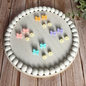 May include: A collection of colorful bow and flower-shaped stud earrings displayed on a round, white beaded tray. The earrings are in shades of pink, yellow, blue, and purple. The tray sits on a wooden surface.