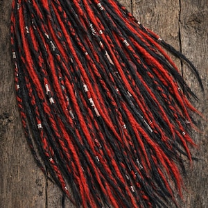 Könnte beinhalten: Ein Bündel aus schwarzen und roten Dreadlocks mit silberfarbenen Metallakzenten. Die Dreadlocks sind auf einer rustikalen Holzoberfläche angeordnet, wodurch die kontrastierenden Farben und Texturen der Haarverlängerungen hervorgehoben werden.