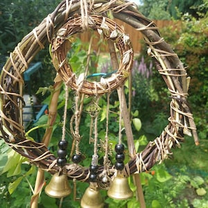 May include: A rustic wind chime with a spiral frame made from woven twigs and twine. It features a smaller circular wreath, brass bells, black beads, and decorative keys hanging from it. The background shows green foliage and a garden setting.