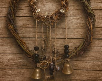 Puede incluir: Una corona rústica hecha a mano con ramitas tejidas, adornada con pequeñas campanas de latón, llaves decorativas y cuentas negras. La corona está suspendida por hilo y colocada sobre un fondo de madera.