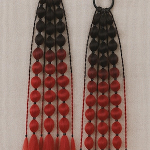 Può includere: Due accessori per capelli con anelli in metallo nero e fili di perline nere e rosse. Le perline passano dal nero al rosso, con nappe rosse in basso. Lo sfondo è un tessuto neutro e strutturato.