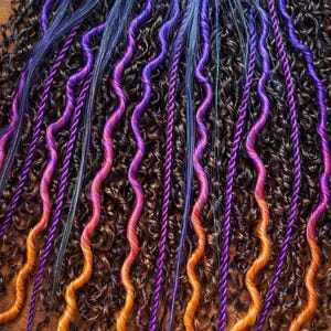 Op de afbeelding: Close-up van krullend bruin haar met levendige paarse, oranje en blauwe accenten. Het haar heeft een mix van texturen, met sommige secties strak opgerold en andere gestyled in golvende strengen. De kleurrijke strengen voegen een speelse en artistieke touch toe.