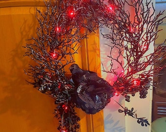May include: A circular wreath constructed from black glittery twigs and branches, adorned with a black artificial bird, dark floral elements, and small dark berries. The wreath is illuminated by a string of small, bright red LED lights.