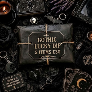 Può includere: Un pacchetto avvolto in carta nera legato con spago, con un'etichetta che recita GOTHIC LUCKY DIP 5 ITEMS £30. Il pacchetto è circondato da vari accessori a tema scuro, tra cui una spilla a forma di teschio, un portachiavi a pipistrello e tarocchi.