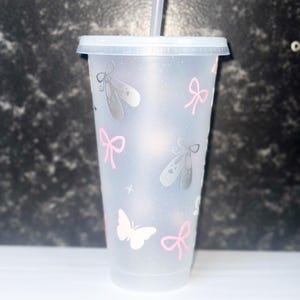 May include: Clear plastic tumbler with a lid and straw, decorated with pink bows, silver ballet slippers, and a white butterfly. The tumbler has a glittery effect.