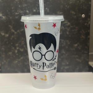 May include: Clear plastic tumbler with a lid and straw, featuring a black silhouette of Harry Potter's face with glasses. The tumbler is decorated with red stars, gold lightning bolts, and the words "Harry Potter".