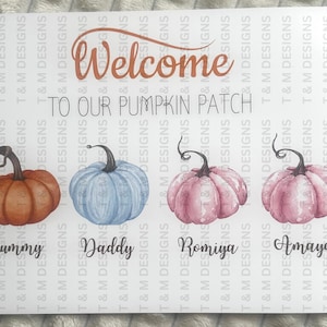May include: A rectangular sign featuring the text Welcome TO OUR PUMPKIN PATCH. Below the text are four watercolour-style pumpkins in orange, light blue, and two shades of pink. Each pumpkin is labelled with the names Mummy, Daddy, Romiya, and Amaya.