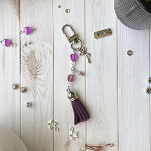 Könnte beinhalten: Ein handgefertigter Schlüsselanhänger mit einem lila Quasten, herzförmigen Perlen und einem silbernen Schleifenanhänger. Der Schlüsselanhänger hat auch ein kleines Metallschild mit der Aufschrift "LOVE". Verstreut sind kleine silberne Perlen und sternförmige Charms.