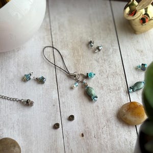 Puede incluir: Un charm de teléfono hecho a mano con un colgante en forma de pez, cuentas azules y blancas y un cordón negro. El charm está sobre una superficie de madera clara con dijes en forma de estrella y una figura de conejo dorado.