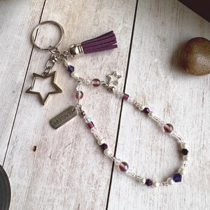Puede incluir: Un llavero plateado con un dije en forma de estrella y una pequeña etiqueta que dice "FEARLESS". El llavero también presenta una borla morada y una cadena de cuentas moradas y transparentes. El fondo es una superficie de madera clara.