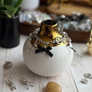 May include: A white and gold ceramic vase with a black bow and silver charm bracelet. The bracelet features musical note and other charms. The vase is decorated with a black bow. Decorative items are scattered around the vase.