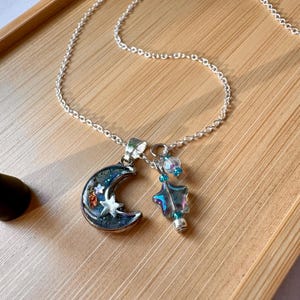 May include: A silver necklace featuring two pendants: a crescent moon and a star-shaped wand. The moon pendant is dark blue with glitter and a small star. The wand pendant has a star shape with iridescent colors and blue beads.