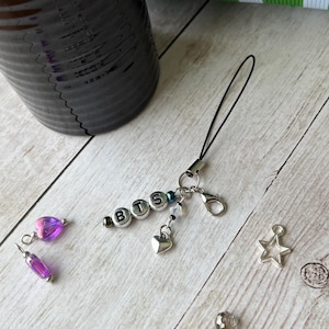 May include: A silver charm with the letters "BTS" spelled out in silver beads. The charm has a black cord and a silver clasp. Additional charms include a heart, a star, and purple beads. The background is a light wood surface.