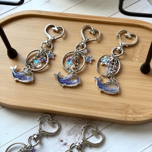 May include: Three whale-shaped keychains with silver-tone hardware and heart-shaped clasps. The whales have a blue and purple galaxy design. Each keychain has a ring with star and crystal charms. The keychains are on a wooden tray.