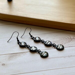 May include: A pair of black drop earrings featuring three circular charms each. The charms contain a dark, glittery background with a white letter H visible on the bottom charm of each earring. The earrings have black metal hooks for attachment.