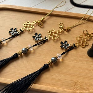 May include: Three decorative charms featuring a gold-toned Chinese knot, a black enamel flower with a central crystal, and a black tassel. Each charm includes a grey pearl bead, a clear faceted bead, and a gold-toned cord for attachment.