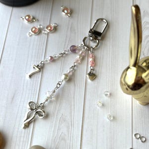 May include: A silver-toned keychain with a lobster clasp, adorned with a bow charm, the number 7, and strands of pink, purple, and clear beads. A small heart charm dangles from the beaded strands. Additional beads and charms are scattered around.