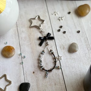 May include: A handmade keychain with a star-shaped clasp and a black bow. The keychain features the word "ECHO" spelled out with letter beads, and is adorned with small beads and a star charm. The color palette includes silver, black, and iridescent beads.