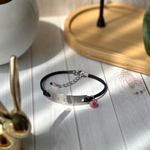Puede incluir: Una pulsera de cordón negro con una placa de metal plateado que dice "BLOOM". La pulsera tiene un cierre de cadena y un pequeño dije de cuentas rosas. También hay una pequeña cuenta rosa adjunta a la pulsera.