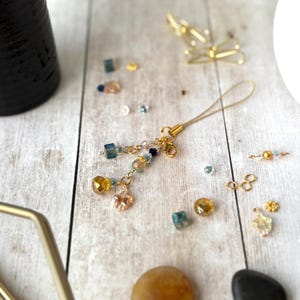 Könnte beinhalten: Ein goldfarbener Anhänger mit Schlaufe, verziert mit hängenden Perlen in Blau-, Gelb- und Pfirsichtönen. Zusätzliche Perlen und kleine goldfarbene Accessoires sind auf einer hellen Holzoberfläche verstreut. Der Anhänger ist wahrscheinlich für ein Telefon oder eine Tasche.