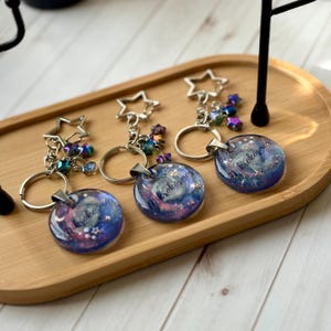 May include: Three keychains with a galaxy design. Each keychain features a round, resin pendant with a swirling blue, purple, and pink galaxy pattern, the word "mikrokosmos", and silver-colored accents. The keychains have silver-colored star-shaped charms and key rings.