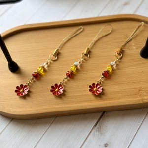 Könnte beinhalten: Drei identische Handy-Anhänger mit einer goldfarbenen Kordel, einer kleinen transparenten Perle, einer gelben Perle, einer dunkelroten Perle und einem roten Emaille-Blumenanhänger mit gelber Mitte. Verbunden durch eine goldfarbene Metallkette.