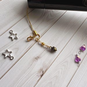 Könnte beinhalten: Ein goldfarbener Telefonanhänger mit Karabinerverschluss, klaren und goldenen Perlen und einem kleinen Eichelanhänger. Zwei silberne Sternanhänger und zwei lila Herzanhänger sind ebenfalls vorhanden. Die Artikel befinden sich auf einer hellen Holzmaserungsoberfläche.
