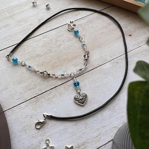 May include: A black cord necklace with a silver chain and a heart pendant. The necklace features blue and clear beads, a star charm, and a lobster clasp. The heart pendant has a decorative openwork design.