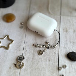 May include: White wireless earbud case with a black lanyard and a charm that spells out "DDAY". The charm includes a silver lotus flower and a pearl. Other silver charms and stones are scattered around the case.